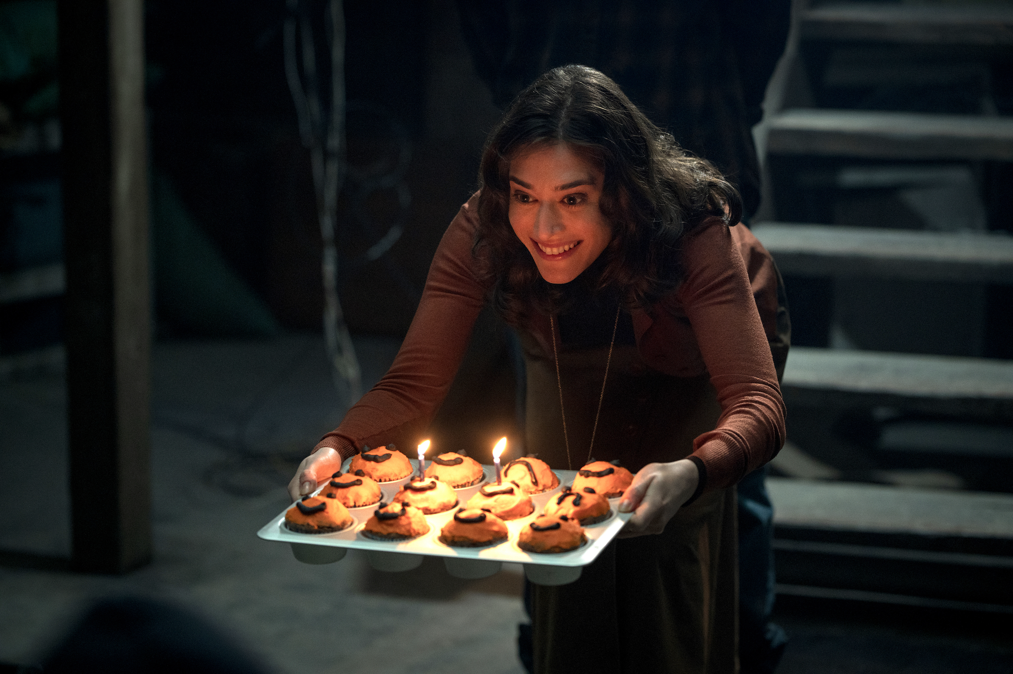 Lizzy Caplan creepily offers a tray of cupcakes, some with small birthday candles, to someone offscreen in the horror movie Cobweb