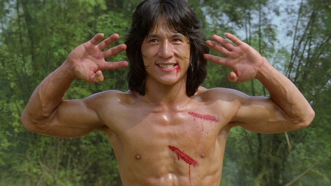 Jackie Chan holds his hands to his face while smiling, shirtless, in Fearless Hyena. He has multiple scratches on his body and bleeds from his mouth.