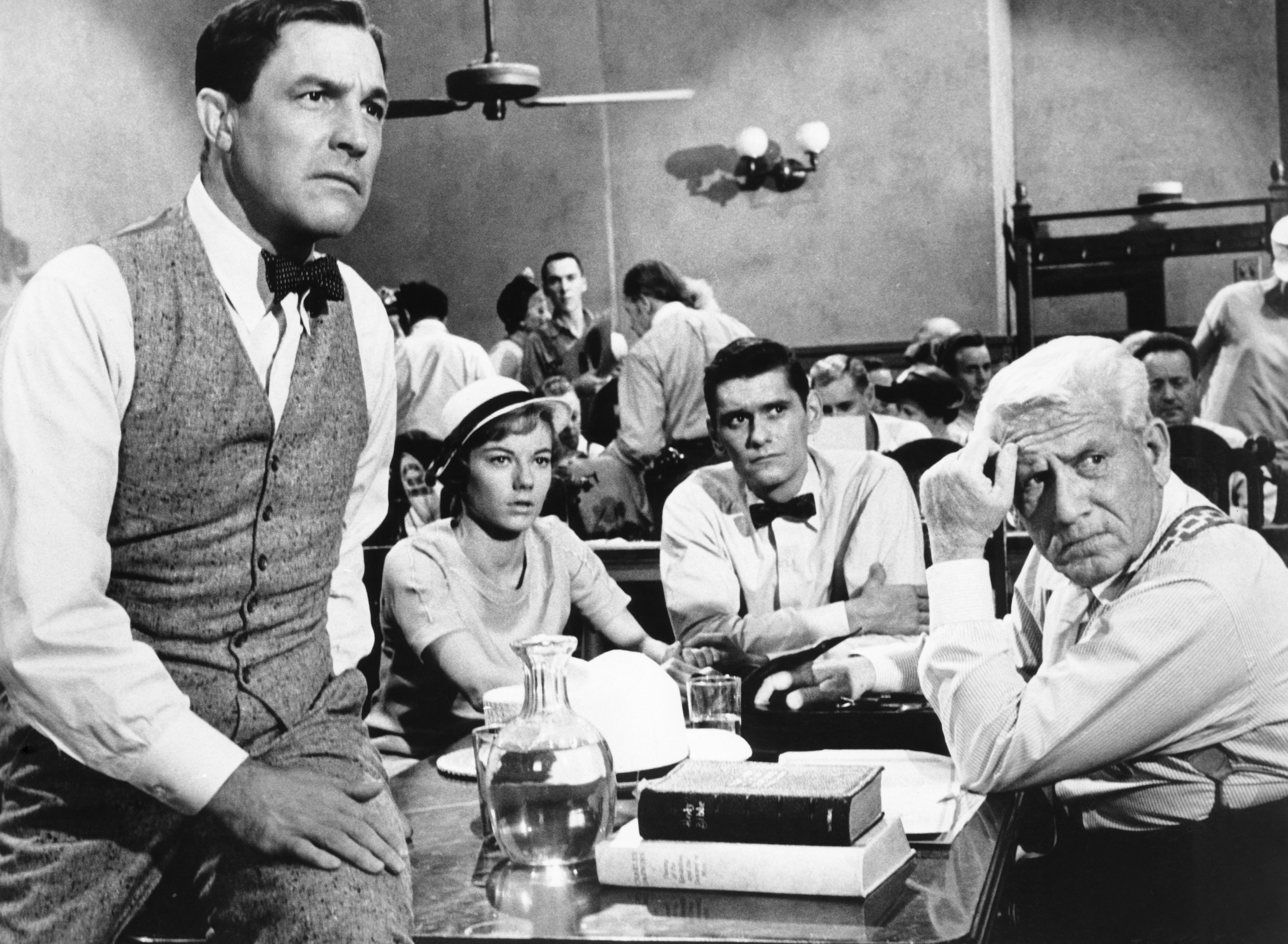 LtR: Gene Kelly as E. K. Hornbeck, Donna Anderson as Rachel Brown, Dick York as Bertram T. Cates, and Spencer Tracy as Henry Drummond in Inherit the Wind. They’re all looking concerned as they sit around the defendant’s table in a packed courtroom (Hornbeck is leaning with one leg up on the table. Brown has a cute little sunhat on and the men are in their shirtsleeves despite the formal setting.