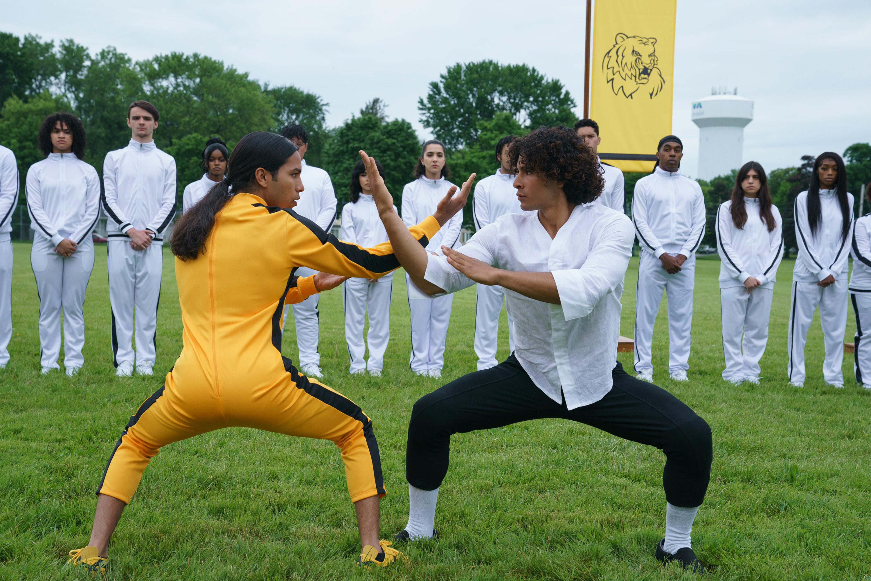 Miguel, dressed in Bruce Lee’s yellow jumpsuit from Game of Death, stands in a martial arts pose at the ready, crossing arms with his opponent in a white shirt and black pants as a row of other teens in white look on in the Hulu Comedy Miguel Wants to Fight